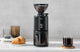 Matte black Eureka Peppe grinder on a kitchen counter beside brewed coffee and a croissant